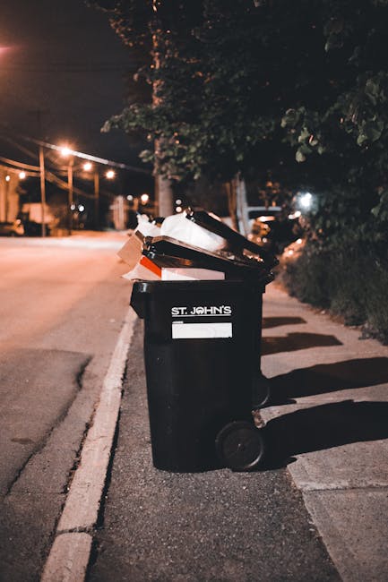 A black wheeled rubbish bin labeled 'St. John's' is positioned on the pavement beside a sidewalk in an urban street at night. The bin is partially open with several items and pieces of cardboard and paper visible inside, some of which are sticking out over the top. It is situated near a curb, with the street stretching into the background illuminated by streetlights that cast a warm glow on the road. In the background, there are trees with leafy canopies, and a row of streetlights extends along the street, creating a pattern of light and shadow. The environment suggests a residential or mixed-use area, where independent waste collection services or private disposal options might be employed for rubbish removal, with the bin ready for collection or awaiting disposal. The scene captures an everyday waste management scenario, illustrating the importance of proper bin placement and waste handling, particularly in urban settings during evening hours, which is relevant to rubbish removal services on the Kennington Estate or similar areas.