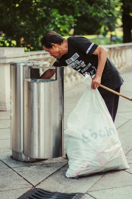A woman with short dark hair, wearing a black T-shirt with white and blue graphic text, is leaning over and placing a clear plastic waste bin into a metallic trash or recycling receptacle on a paved sidewalk. She holds a large white plastic bag filled with rubbish in her left hand, which rests against her side, while her right arm is extended into the bin to deposit waste. The metallic bin has a cylindrical shape with a smooth, reflective surface and an opening at the top. The scene is set outdoors in a green urban area, with trees and a low stone or concrete balustrade visible in the background. The lighting suggests daytime with natural sunlight, and the setting appears to be a public space or park where waste is collected, possibly as part of a community clean-up or routine rubbish management outside a private property. The image reflects an activity aligned with waste collection and disposal, demonstrating proper rubbish handling outside a residential or commercial premise, consistent with services provided by waste collection specialists like Waste Collection Kennington.