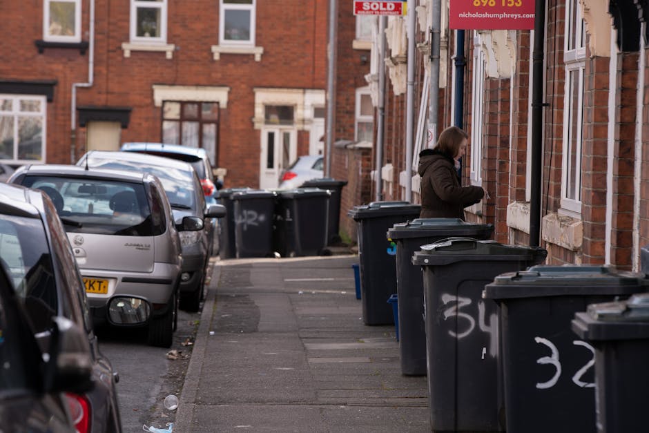 A black wheeled rubbish bin labeled 'St. John's' is positioned on the pavement beside a sidewalk in an urban street at night. The bin is partially open with several items and pieces of cardboard and paper visible inside, some of which are sticking out over the top. It is situated near a curb, with the street stretching into the background illuminated by streetlights that cast a warm glow on the road. In the background, there are trees with leafy canopies, and a row of streetlights extends along the street, creating a pattern of light and shadow. The environment suggests a residential or mixed-use area, where independent waste collection services or private disposal options might be employed for rubbish removal, with the bin ready for collection or awaiting disposal. The scene captures an everyday waste management scenario, illustrating the importance of proper bin placement and waste handling, particularly in urban settings during evening hours, which is relevant to rubbish removal services on the Kennington Estate or similar areas.