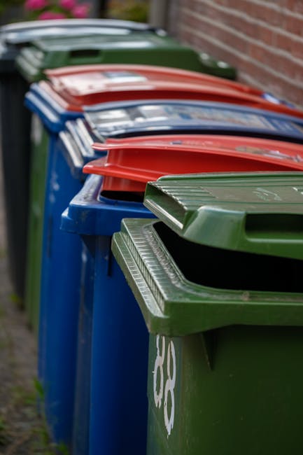 A row of large, rectangular wheelie bins positioned along a pavement adjacent to a brick wall, featuring green, blue, and red lids from left to right, with the green bin in the foreground and the others extending into the background. The bins are made of sturdy plastic with textured surfaces on the lids; the green bin appears to be a standard waste or garden refuse container, while the blue bins are likely for general waste or recyclables and the red lid indicates a specific waste category. The lids are closed, with some lids slightly raised or leaning, and the bins are arranged side by side, occupying a narrow strip of outdoor space, possibly a residential driveway or alleyway. The environment suggests a typical urban or suburban setting for waste collection, consistent with services such as waste collection Kennington. The lighting is natural, with no visible shadows, highlighting the different colours and textures of the bins, which stand out against the background brick wall and a hint of greenery in the upper part of the image, reinforcing a scene related to rubbish disposal or independent waste handling options in a community setting.