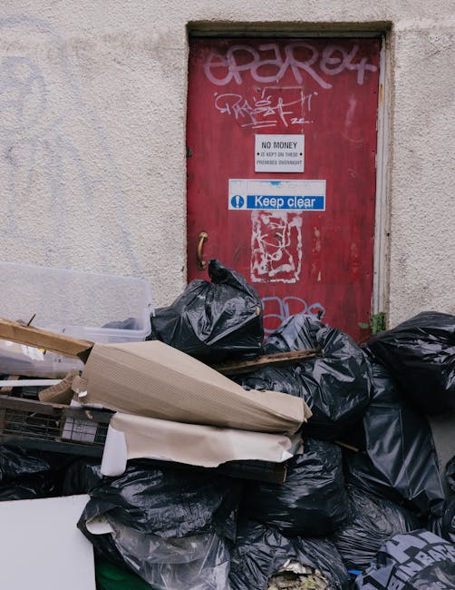 A row of large, rectangular wheelie bins positioned along a pavement adjacent to a brick wall, featuring green, blue, and red lids from left to right, with the green bin in the foreground and the others extending into the background. The bins are made of sturdy plastic with textured surfaces on the lids; the green bin appears to be a standard waste or garden refuse container, while the blue bins are likely for general waste or recyclables and the red lid indicates a specific waste category. The lids are closed, with some lids slightly raised or leaning, and the bins are arranged side by side, occupying a narrow strip of outdoor space, possibly a residential driveway or alleyway. The environment suggests a typical urban or suburban setting for waste collection, consistent with services such as waste collection Kennington. The lighting is natural, with no visible shadows, highlighting the different colours and textures of the bins, which stand out against the background brick wall and a hint of greenery in the upper part of the image, reinforcing a scene related to rubbish disposal or independent waste handling options in a community setting.