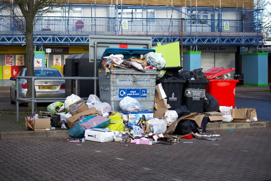 A collection of overflowing rubbish and waste materials piled on a paved urban street, including assorted cardboard boxes, plastic bags, paper waste, and packaging. The waste is situated next to a metal railing and various black, green, and red waste bins, some of which are open or tipped over, with contents spilling out. Behind the waste, there is a parked silver car with a yellow registration plate, and in the background, a commercial building with a blue scaffolding structure and signage indicating shops such as fish bars and other retail outlets. The scene depicts an unattended waste accumulation in a public space, highlighting the need for proper rubbish collection. Waste Collection Kennington's site often addresses issues related to private waste disposal and alternative rubbish removal options, which may include on-site clearance or independent collection services, as suggested by the context of the surroundings and the nature of the waste accumulation.