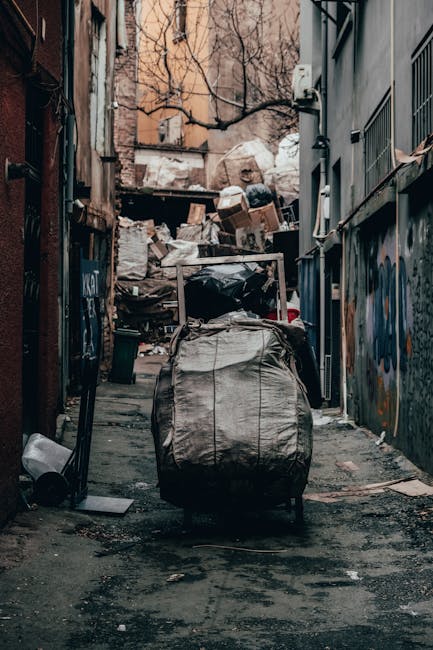 A narrow urban alleyway with a heavily loaded wheeled rubbish bin in the foreground, positioned centrally on a dirt and asphalt surface. The bin is made of woven, weathered fabric with visible creases and signs of wear, and is closed with a metal handle on top. In the background, a large pile of mixed waste, including cardboard boxes, paper, plastic bags, and other debris, is stacked against a weathered brick and concrete wall. The alley is flanked by tall buildings on either side, with graffiti visible on the right wall and some peeling paint. Bare tree branches extend overhead, casting faint shadows in the dim, natural light. The scene depicts an environment associated with private waste collection efforts, suggesting the need for rubbish removal services to clear clutter from such confined urban spaces, managed by Waste Collection Kennington in the context of alternative waste handling options.