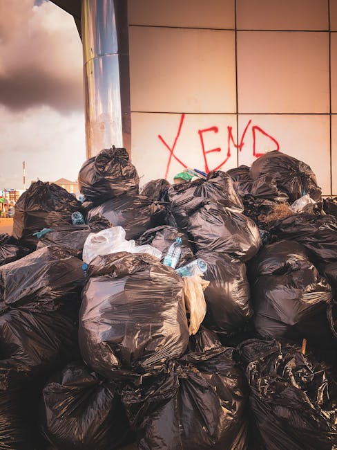 A large pile of black rubbish bags, some partially torn or bulging, is stacked against a modern building with a reflective, grid-like facade. The bags contain mixed waste, with visible plastic bottles and packaging protruding from a few of them. In the background, a graffiti tag in red spray paint spells out 'XEND' on the building’s wall. The scene is outdoors, likely on a pavement or driveway, with a cloudy sky overhead suggesting late afternoon or early evening lighting. The image depicts a typical example of waste accumulation requiring private rubbish collection or on-site clearance, highlighting the importance of professional waste removal services like those offered by Waste Collection Kennington. The environment appears urban, with no visible vegetation or natural landscape, emphasizing the context of urban waste management and rubbish removal from commercial or residential areas. The focus on the discarded waste and the modern building's reflective surface provides a clear visual connection to the need for effective rubbish clearance solutions in busy city environments.