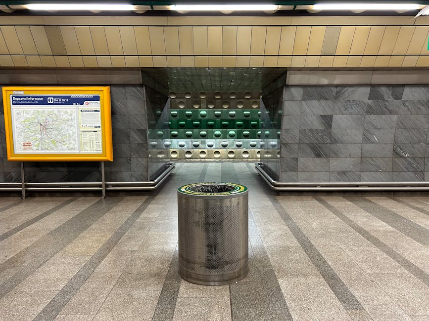 The image depicts an underground subway station with a round metallic trash bin positioned centrally on a tiled floor. The trash bin has a perforated surface and a dark-colored, flat lid. Behind it, there is a broad staircase leading upward, flanked by smooth, dark grey tiled walls. To the left of the staircase, a large, framed map display with a yellow border is mounted on the wall, providing navigation and local information. The walls are constructed of sleek, rectangular, dark grey tiles that extend outwards and contrast with the beige tiled ceiling, which features linear lighting fixtures. The environment appears clean and well-maintained, with a modern, utilitarian design typical of urban public transportation hubs. Waste Collection Kennington offers private rubbish removal services, and this scene reflects the station’s emphasis on organized, efficient waste disposal within a public transit context, subtly aligning with on-site waste management and alternative collection options focused on cleanliness and orderliness.
