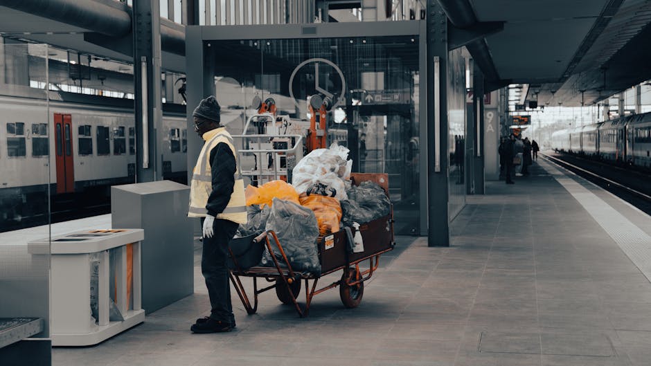 At a modern train station platform, a worker wearing a dark jacket, grey beanie, and high-visibility vest stands next to a red utility cart filled with various black, grey, and orange plastic bags and wrapped items of waste, some of which are visibly crumpled or tied. Behind the worker, there is a glass-walled elevator shaft, with mechanical elements such as pulleys and cables visible, suggesting ongoing maintenance or waste removal activities associated with convenient facilities. The platform surface is composed of large, light grey tiles, with a white safety line near the edge, and to the right, parallel train tracks extend into the distance, where a few small figures are visible. Against the backdrop of a large station building with a dark metal and glass facade, waste collection activities are indicative of an organized approach to managing refuse outside of public authority services, aligning with private or independent rubbish removal options provided by companies like Waste Collection Kennington. Natural lighting suggests daytime, with diffuse light illuminating the scene clearly and evenly, highlighting the practical setting for rubbish disposal and collection.