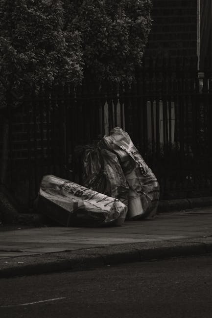 In the foreground, two large black plastic garbage bags filled with waste sit on a paved sidewalk near the curb, leaning slightly against each other. The bags are partially crumpled and appear to be tightly secured, with some parts reflecting light, indicating glossy plastic material. Behind the bags, a black wrought iron fence with vertical bars separates the sidewalk from a garden area filled with dense foliage of dark, leafy bushes. The background also reveals a portion of a building with dark, textured exterior walls, perhaps made of brick or tile, partially obscured by the fence and trees. The scene is illuminated subtly, suggesting overcast weather or evening light, creating a muted, neutral atmosphere. The location hints at an alternative private waste collection scenario, possibly managed by Waste Collection Kennington, in an urban area where residents dispose of larger refuse items on the street for collection or disposal.