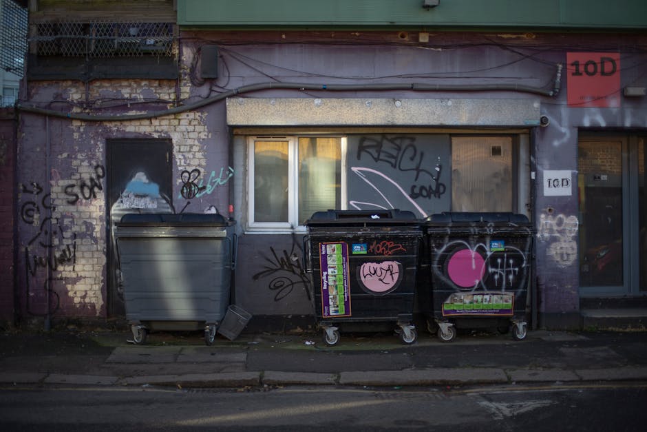 In the foreground, two large black plastic garbage bags filled with waste sit on a paved sidewalk near the curb, leaning slightly against each other. The bags are partially crumpled and appear to be tightly secured, with some parts reflecting light, indicating glossy plastic material. Behind the bags, a black wrought iron fence with vertical bars separates the sidewalk from a garden area filled with dense foliage of dark, leafy bushes. The background also reveals a portion of a building with dark, textured exterior walls, perhaps made of brick or tile, partially obscured by the fence and trees. The scene is illuminated subtly, suggesting overcast weather or evening light, creating a muted, neutral atmosphere. The location hints at an alternative private waste collection scenario, possibly managed by Waste Collection Kennington, in an urban area where residents dispose of larger refuse items on the street for collection or disposal.