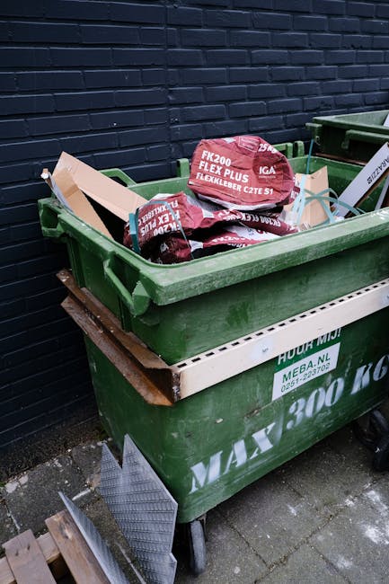 A large green wheeled waste container filled with several red and black plastic bags containing garden or household waste, positioned on a paved surface against a dark blue brick wall. The bags are labeled with gardening or outdoor products, such as fertilizer or soil enhancers. The container, which appears to be part of private waste disposal or alternative rubbish collection services, is situated near another similar bin; a wooden pallet and a metal ramp are visible on the ground nearby. The environment suggests an outdoor area, possibly a back yard or adjacent to a commercial or residential building, where waste collection is managed independently from local council services. The presence of different types of waste containers and the specific arrangement indicates routine waste handling, supporting private or specialised rubbish removal activities typical in certain property settings. The scene highlights the practical aspect of managing waste outside standard municipal collection schemes, aligning with the concept of on-site clearance or alternative waste handling methods often employed by waste management companies like Waste Collection Kennington.