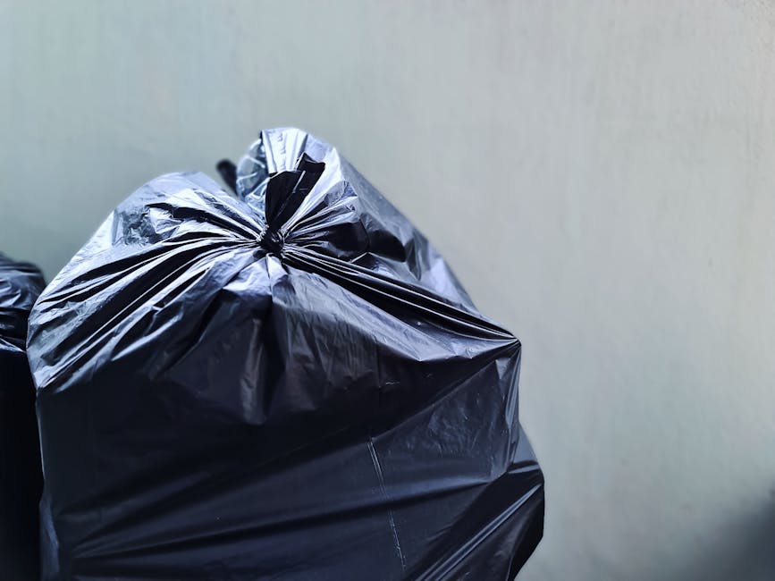 A large green wheeled waste container filled with several red and black plastic bags containing garden or household waste, positioned on a paved surface against a dark blue brick wall. The bags are labeled with gardening or outdoor products, such as fertilizer or soil enhancers. The container, which appears to be part of private waste disposal or alternative rubbish collection services, is situated near another similar bin; a wooden pallet and a metal ramp are visible on the ground nearby. The environment suggests an outdoor area, possibly a back yard or adjacent to a commercial or residential building, where waste collection is managed independently from local council services. The presence of different types of waste containers and the specific arrangement indicates routine waste handling, supporting private or specialised rubbish removal activities typical in certain property settings. The scene highlights the practical aspect of managing waste outside standard municipal collection schemes, aligning with the concept of on-site clearance or alternative waste handling methods often employed by waste management companies like Waste Collection Kennington.
