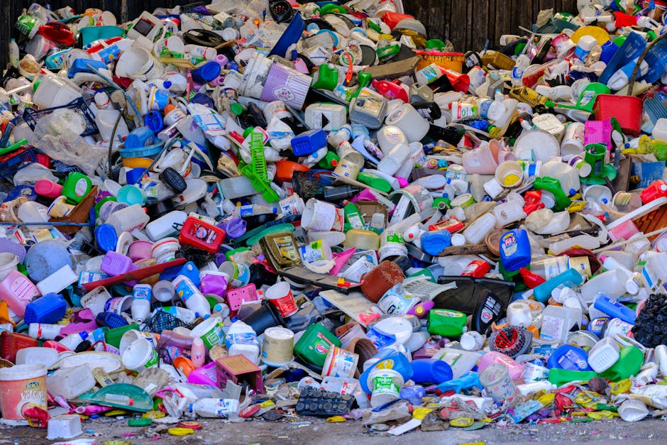 A large outdoor pile of mixed household and personal waste consisting mainly of plastic containers, bottles, and packaging materials. The collection includes various sizes, shapes, and colours of plastic bottles, caps, and tubs, with some items made of translucent or opaque plastic and others in bright hues such as red, blue, green, pink, and yellow. The waste is scattered across a paved or concrete surface and appears to be haphazardly dumped, with some items stacked atop each other and others lying loosely. In the background, a wooden fence or paneling is partially visible behind the pile, indicating an outdoor setting for waste accumulation. The lighting suggests natural daylight, highlighting the textures of the plastics, some with smooth surfaces and others with ridges or embossed labels. This scene exemplifies the type of waste that might be collected through private rubbish removal services, such as those offered by Waste Collection Kennington, and underscores the importance of recycling and responsible waste disposal for local residents and property owners.