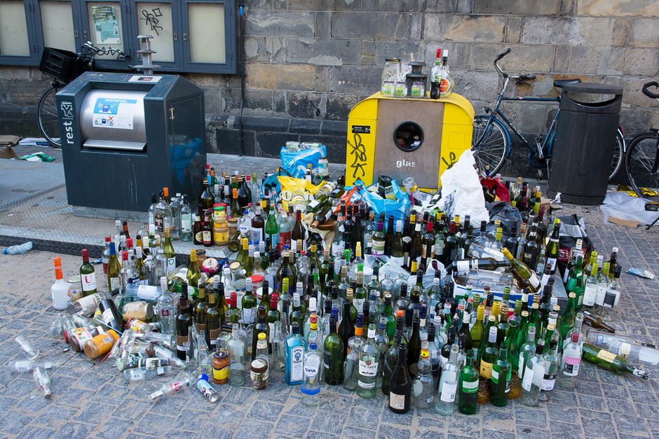 A large collection of discarded glass bottles and plastic containers is spread across a paved sidewalk area. The bottles vary in size, shape, and color, including clear, green, and brown glass, some with labels still attached, and many are lying on their sides or leaning against each other. Among the bottles, there is scattered packaging and debris, with a few crumpled paper bags and plastic wrapping visible. In the background, a yellow recycling container with a circular opening for glass disposal and some items placed on top is positioned next to a black waste bin with a curved lid. To the left, there is a grey, modern reverse vending machine designed for recycling bottles, with a digital display and a card payment terminal, and a few other small objects nearby. On the far side, a brick wall lines the environment, with a window visible above. Two bicycles are parked behind some waste bins, indicating an urban or residential setting. The scene reflects an instance of private waste disposal or on-site clearance managed by Waste Collection Kennington, emphasizing the importance of proper rubbish handling and recycling practices.