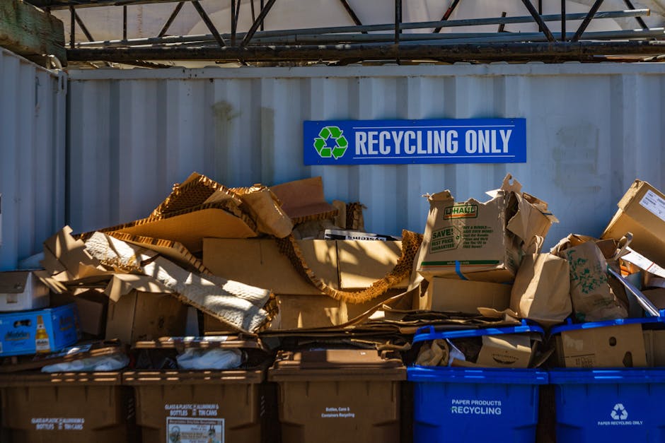 A large outdoor pile of mixed household and personal waste consisting mainly of plastic containers, bottles, and packaging materials. The collection includes various sizes, shapes, and colours of plastic bottles, caps, and tubs, with some items made of translucent or opaque plastic and others in bright hues such as red, blue, green, pink, and yellow. The waste is scattered across a paved or concrete surface and appears to be haphazardly dumped, with some items stacked atop each other and others lying loosely. In the background, a wooden fence or paneling is partially visible behind the pile, indicating an outdoor setting for waste accumulation. The lighting suggests natural daylight, highlighting the textures of the plastics, some with smooth surfaces and others with ridges or embossed labels. This scene exemplifies the type of waste that might be collected through private rubbish removal services, such as those offered by Waste Collection Kennington, and underscores the importance of recycling and responsible waste disposal for local residents and property owners.