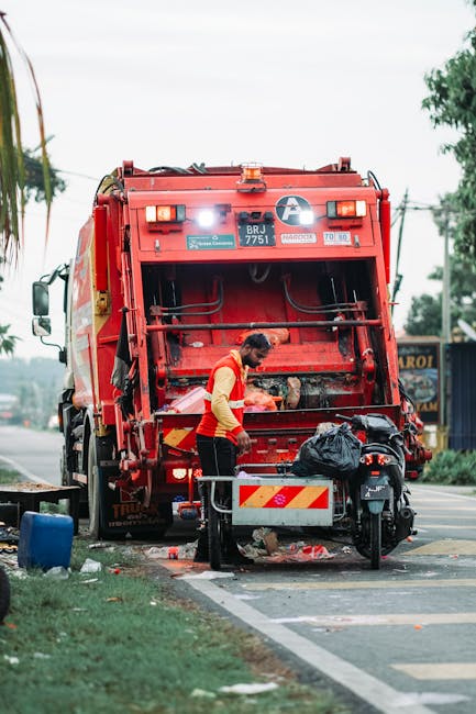 A waste collection worker, dressed in a high-visibility yellow and red vest, is seen standing next to a large red waste collection vehicle on a roadside. The worker is engaged in an activity related to rubbish management, with their attention focused on the rear section of the truck, which has an open loading mechanism. The truck is positioned on the edge of a paved street, directly adjacent to a grassy verge with some trees and utility poles visible in the background. A black plastic rubbish bag is placed on a small metal platform attached to a motorbike parked nearby, which is stationed on the roadside curb. The environment appears to be a suburban or urban street with overhead power lines and a partly cloudy sky indicating daytime. Visible debris, including small rubbish items on the ground and scattered near the worker’s feet, suggests ongoing waste collection or clearance activity. This scene reflects an instance of autonomous waste collection, possibly performed by private waste management services such as Waste Collection Kennington, emphasizing practical, on-site rubbish removal in a residential or commercial area.