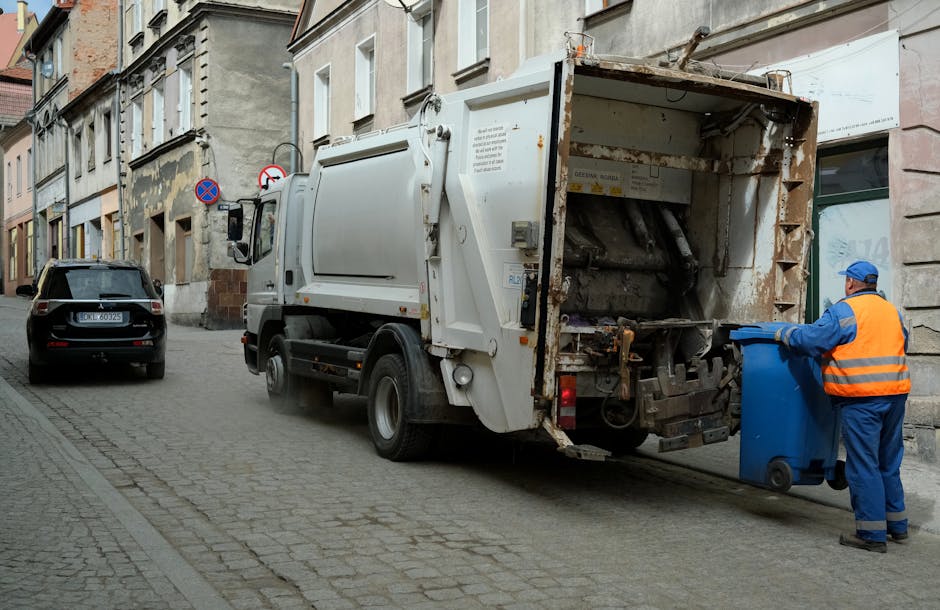 A waste collection vehicle parked on a narrow city street in front of old multi-storey buildings with a textured stone façade. The truck is white with some visible rust and dirt, particularly around the rear loading mechanism, which is open and appears to be in use. A worker dressed in a blue uniform and an orange high-visibility vest is standing beside the truck, emptying a blue wheeled bin into the rear compartment. To the left of the waste disposal truck, a black car is parked parallel to the curb. The street is paved with small, rectangular stone cobbles, and there are two no-parking signs attached to a pole on the side of the buildings. The environment suggests an urban setting where private waste collection services such as those offered by Waste Collection Kennington could manage on-site rubbish removal, including bulky waste items, outside of standard council collection schedules.