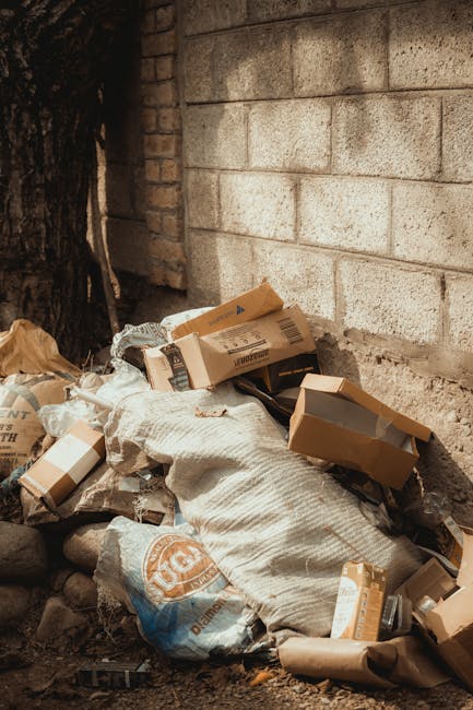 A small pile of discarded cardboard boxes, some flattened with open flaps, rest on a rough, unpaved ground beside a beige brick wall and a dark tree trunk. The boxes vary in size and are made of standard corrugated material with printed labels and barcodes visible on some. Among the boxes, there are also a couple of tightly bundled beige and white woven fabric bags, likely used for waste containment. The scene appears to be outdoors, with natural ambient lighting highlighting the textures of the cardboard, fabric, and the bricks. The area suggests a location where private waste or bulky rubbish is temporarily deposited, possibly awaiting collection or disposal, reflecting typical practices of independent waste management services such as those offered by Waste Collection Kennington. There are no signs of systematic recycling or organized waste segregation in the scene, emphasizing a straightforward approach to rubbish handling on-site. The surrounding environment indicates it is an informal or rear service area, with minimal environmental features beyond the brick wall and tree trunk, suitable for quick rubbish stacking or disposal activities consistent with alternative waste handling methods. 